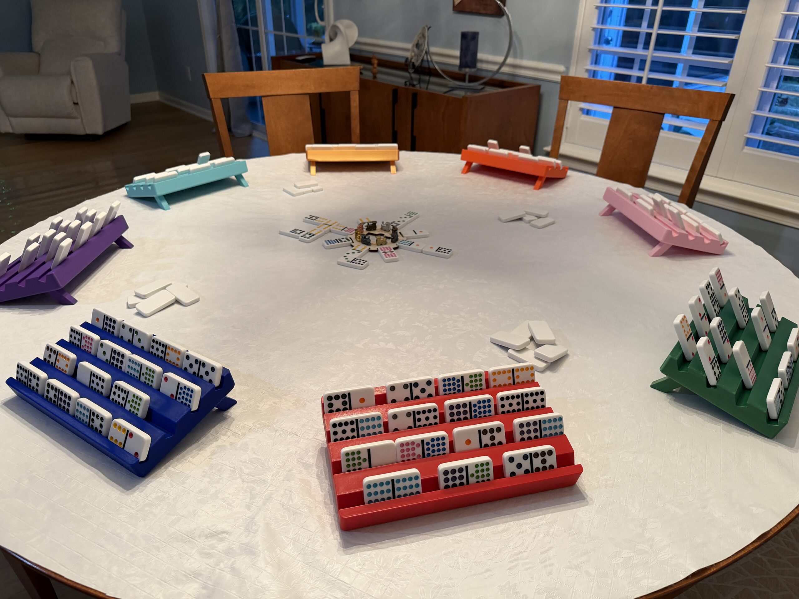 Custom 3D-printed domino racks in multiple colors arranged around a dining table during a multiplayer domino game.